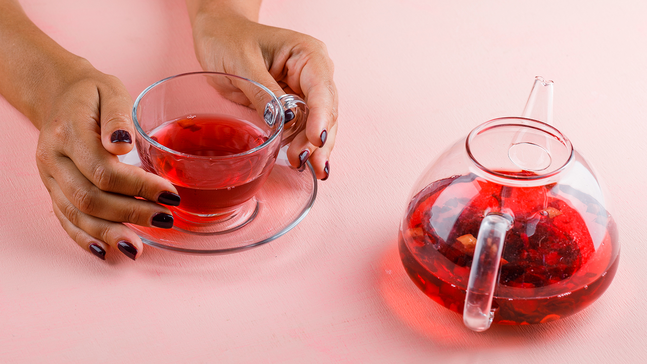 A woman holding a cup of hibiscus herbal tea next to a clear teapot 
